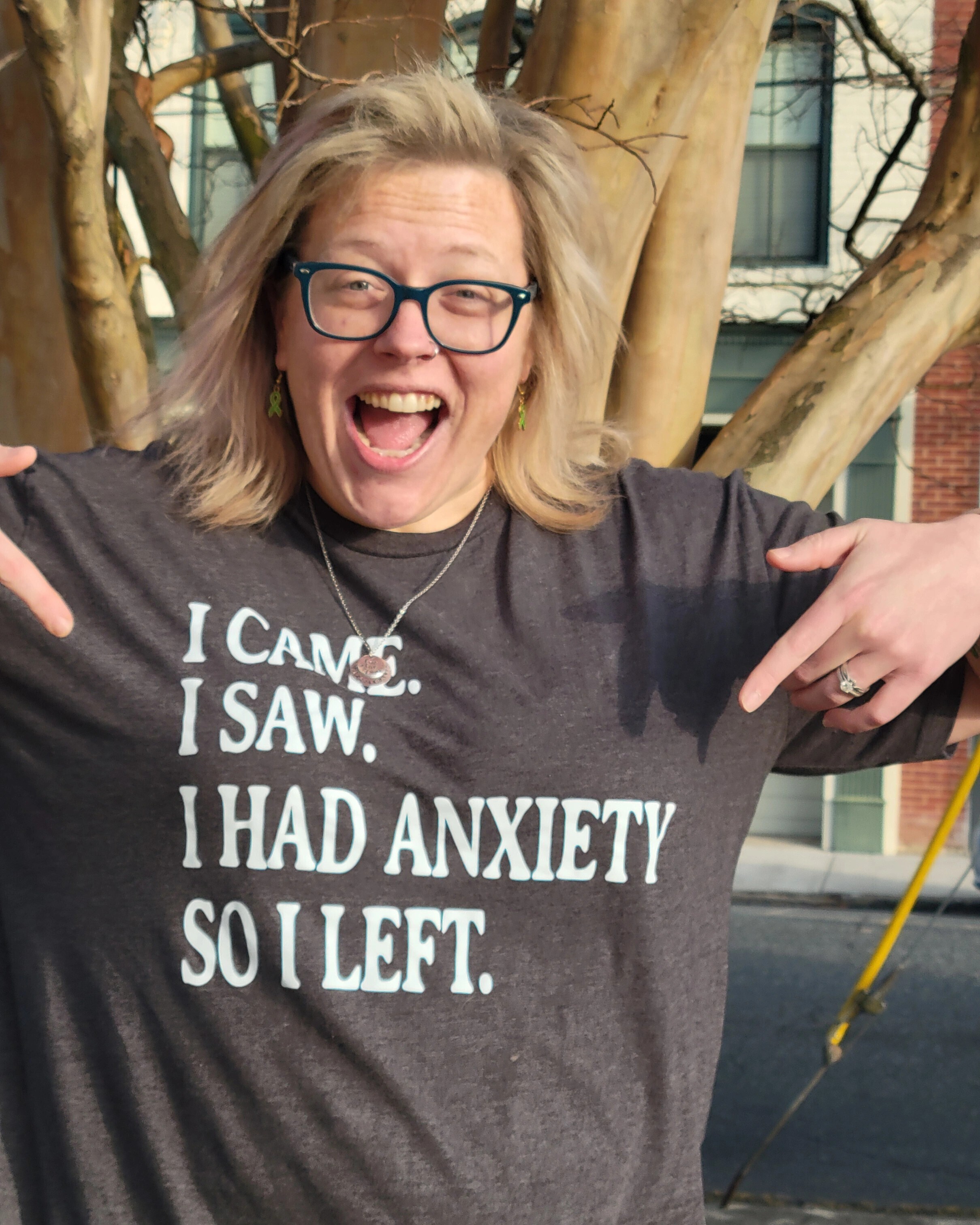 Smiling woman pointing to her T-shirt that reads “I came. I saw. I had anxiety so I left.” promoting mental health awareness with humor