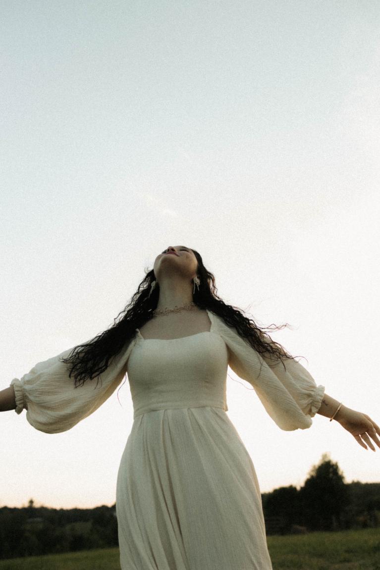 Girl with long brown hair in white maxi dress standing outside with her arms outstretched and looking at the sky