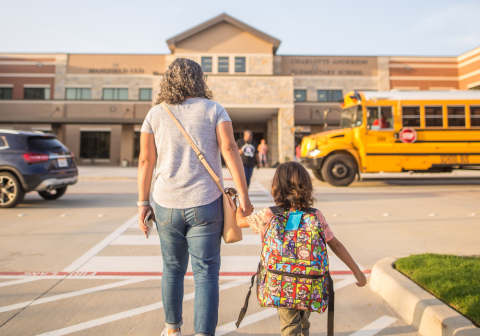 A woman walking a child to school, a bus in the parking lot in front of the school