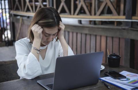  Woman sitting at a table looking at her laptop and fisting her hands against her head due to stress