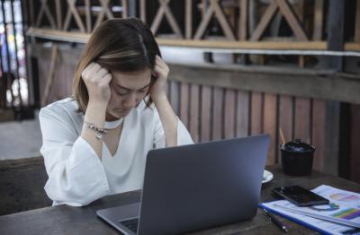  Woman sitting at a table looking at her laptop and fisting her hands against her head due to stress
