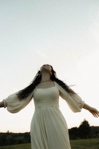 Girl with long brown hair in white maxi dress standing outside with her arms outstretched and looking at the sky