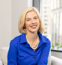 Professional headshot of a woman with blonde hair, wearing a navy blue blouse and a pearl necklace, smiling warmly at the camera.