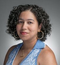 A woman with curly hair wearing a blue sleeveless top, gold hoop earrings, and a delicate necklace. She is smiling warmly against a neutral gray background
