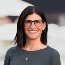 Professional headshot of a woman with dark hair, wearing glasses and a black top, smiling warmly at the camera.